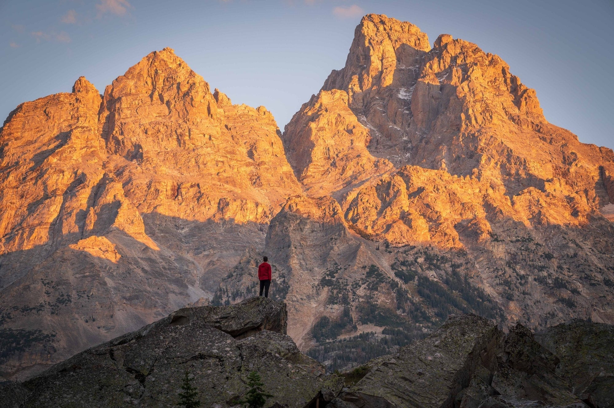Sunrise light hitting the Cathedral Group in Grand Teton National Park with a lone hiker standing on a rocky foreground ridge.