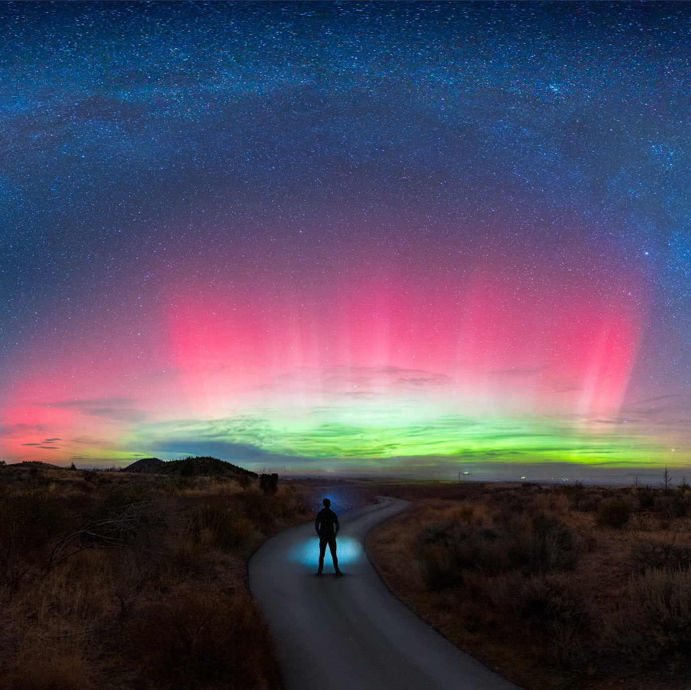 Silhouetted hiker walking along desert road under vivid aurora borealis and Milky Way in Lava Beds National Monument