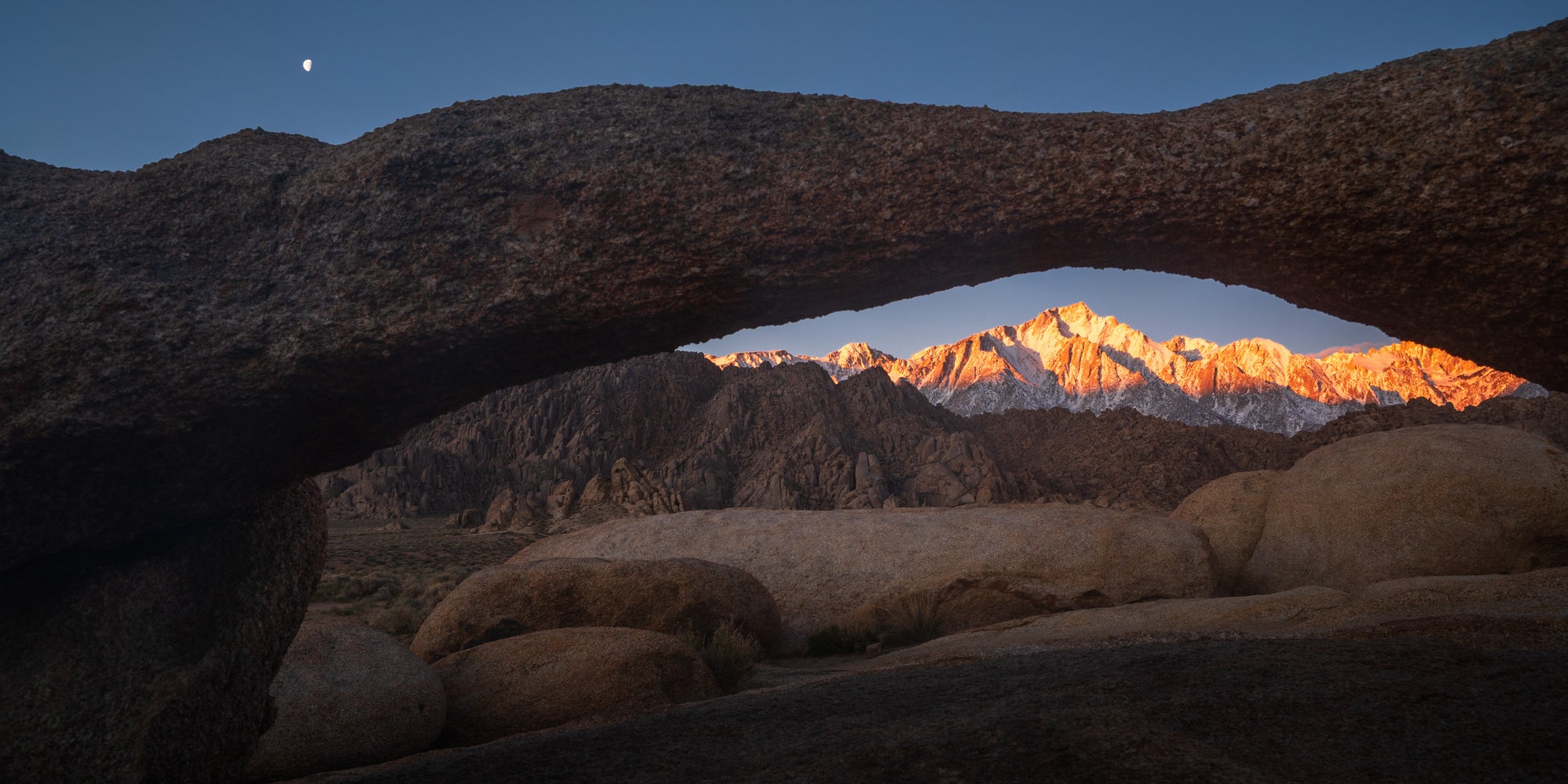 Sunrise view of Lone Pine Peak framed by a natural arch in the Alabama Hills, California, with warm light on the Sierra crest and a moon in the early sky.