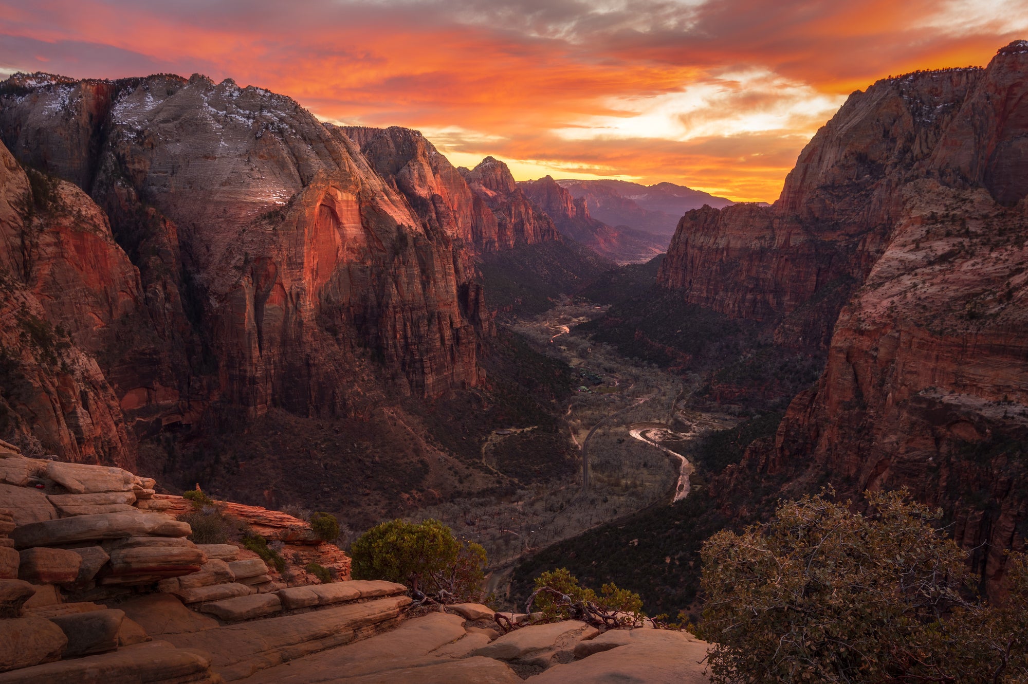 Fiery sunset over Zion Canyon viewed from the summit of Angels Landing, with red sandstone cliffs and the Virgin River winding through the canyon below.