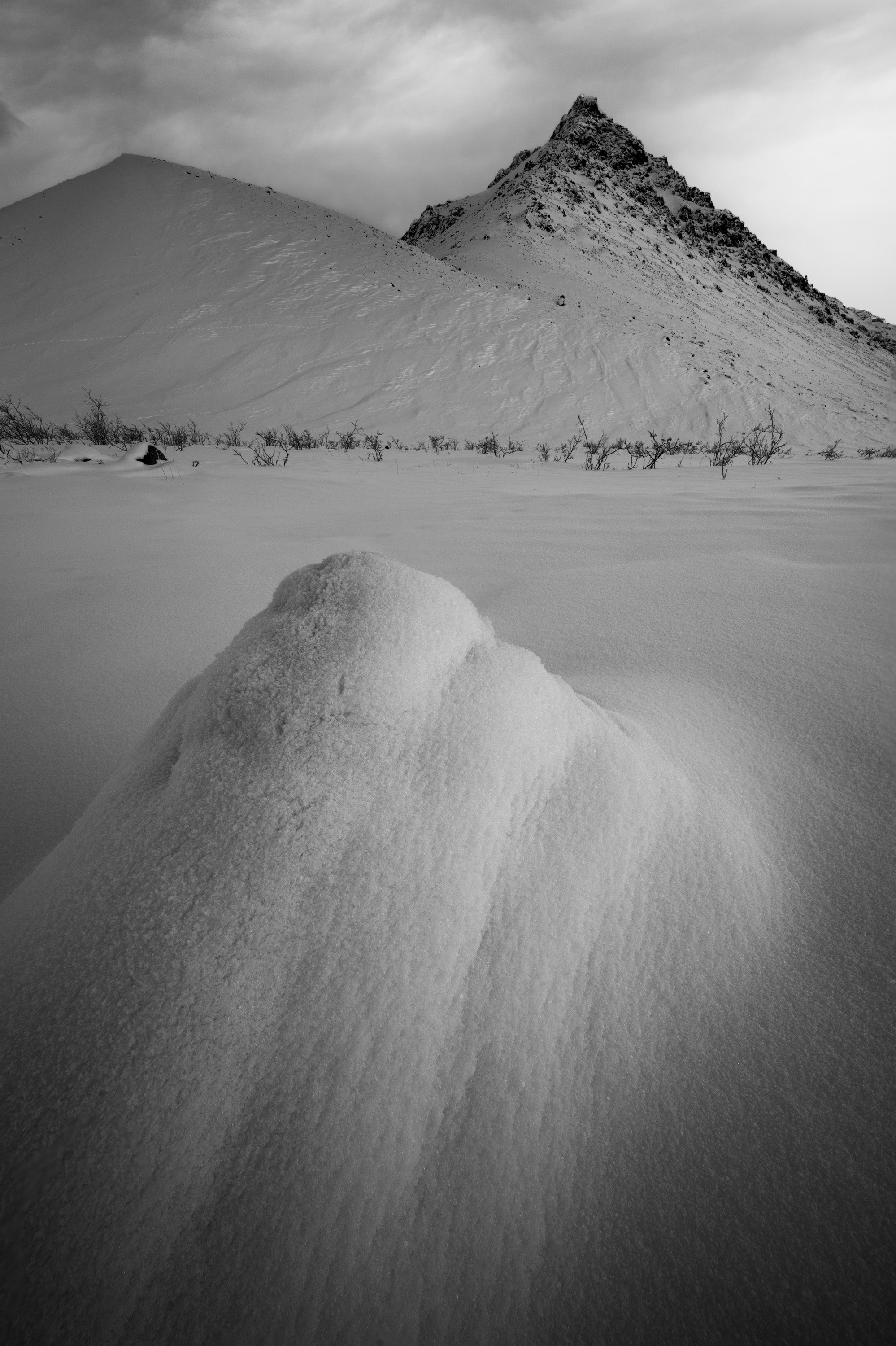 Black-and-white winter landscape on Alaska’s North Slope near the Arctic National Wildlife Refuge, showing a snow mound mirroring the shape of a distant triangular mountain.