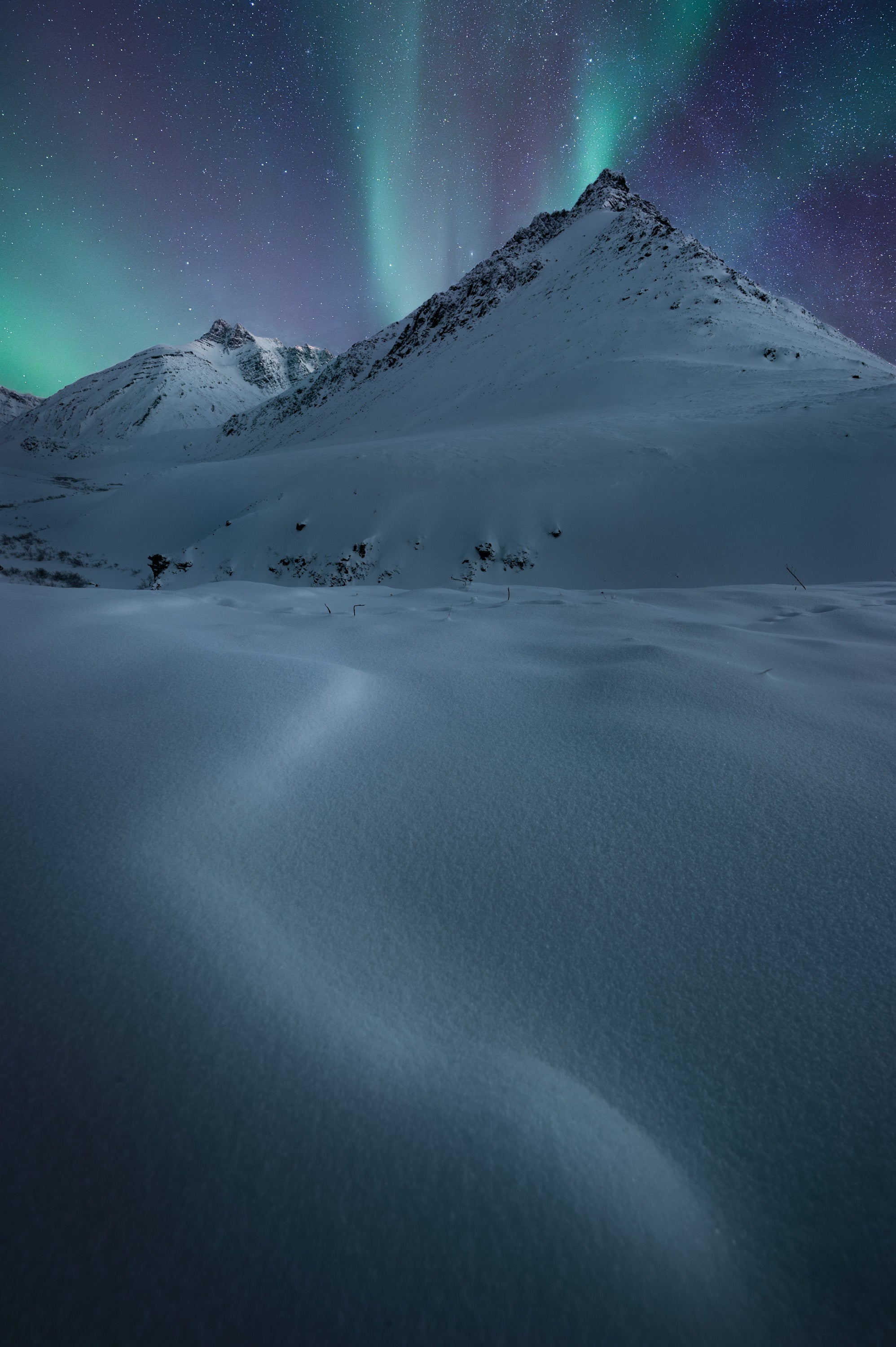 Aurora borealis above a snow-covered mountain in Alaska’s Arctic National Wildlife Refuge, with wind-shaped snowdrifts in the foreground under a clear winter night sky.