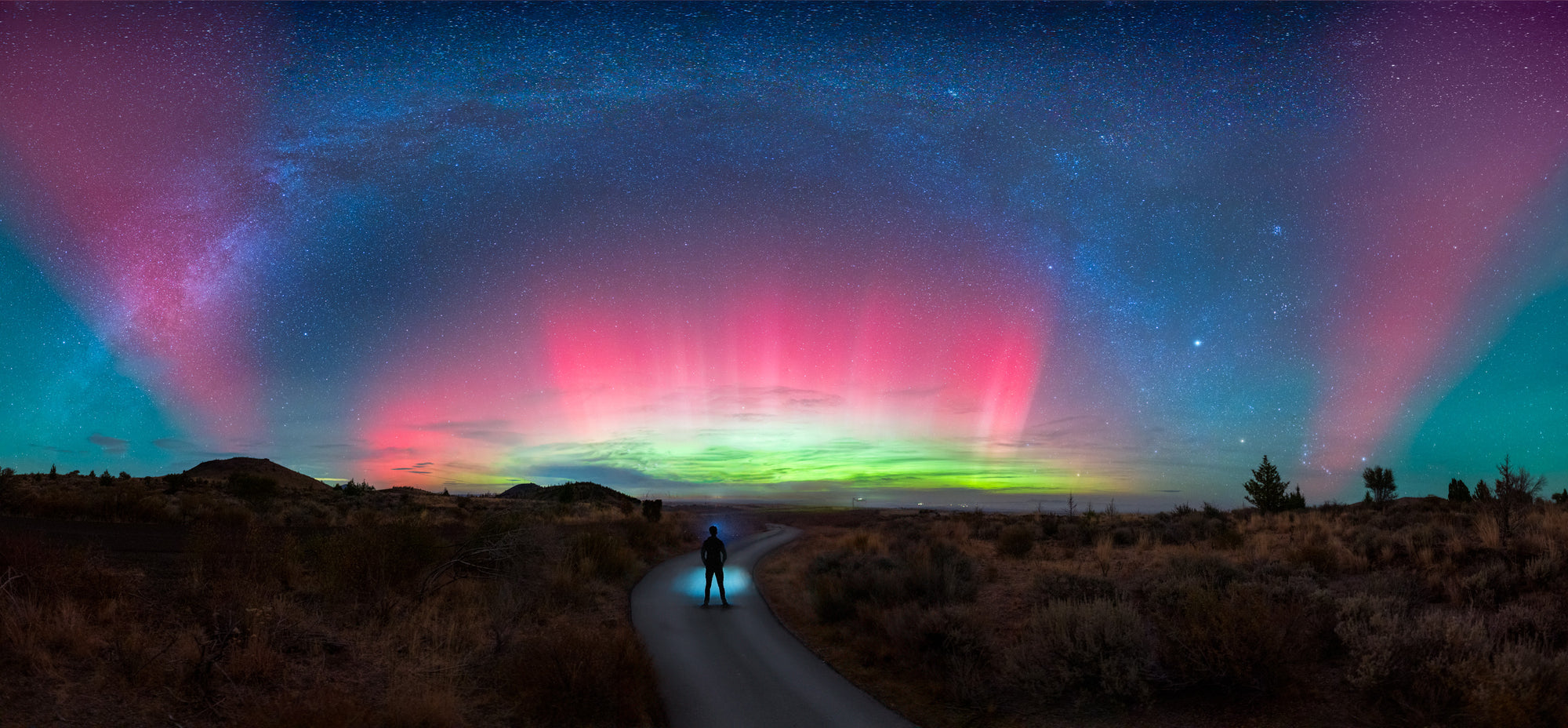 Man walking on winding dirt road in Lava Beds National Monument under vivid aurora borealis and faint Milky Way at night
