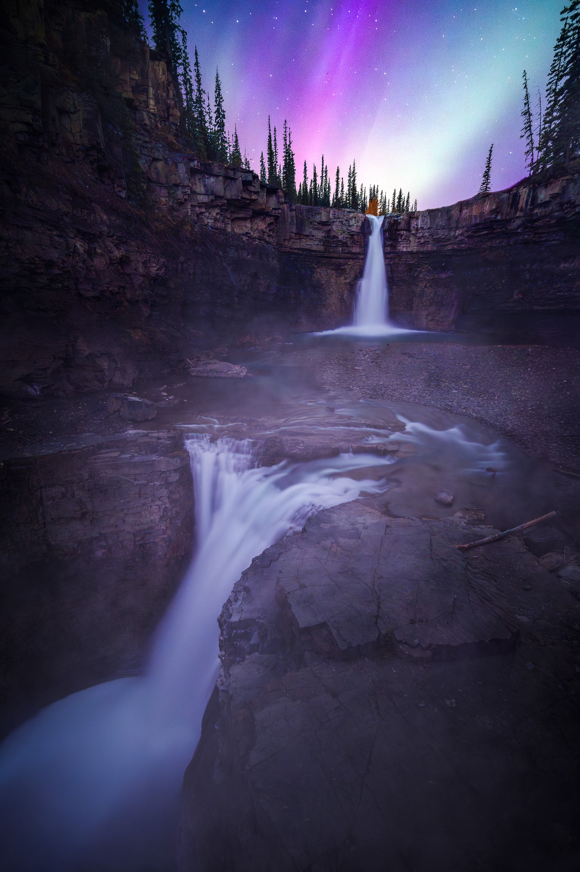 Aurora borealis (Northern Lights) glowing above a powerful multi-tier waterfall in Alberta’s Kootenay Plains, with cascading water flowing through a rocky canyon at night.
