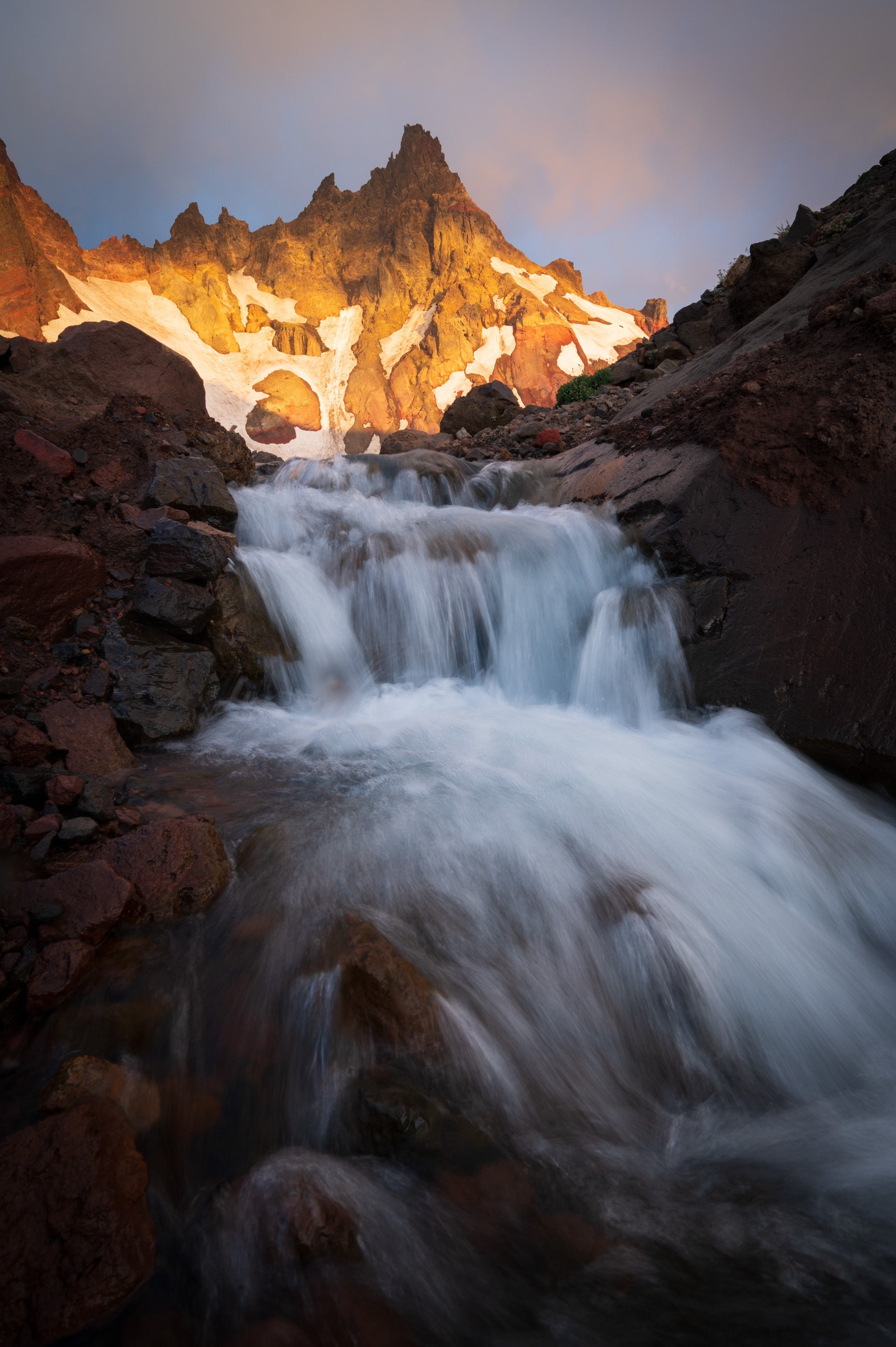 Sunrise light on Broken Top volcano in the central Oregon Cascades, with a small waterfall flowing through dark volcanic rock beneath jagged peaks and lingering snow.