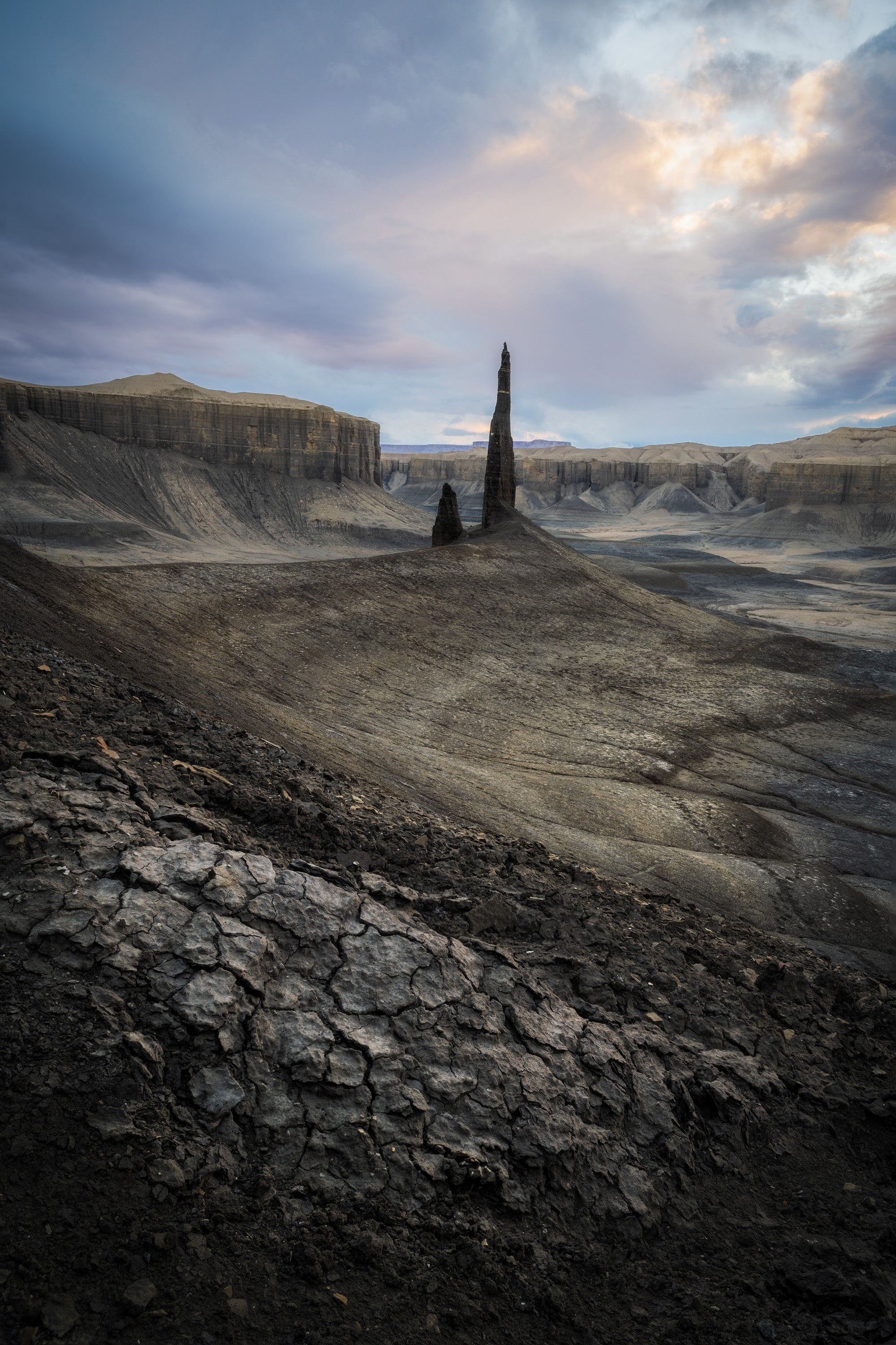 Tall rock spire rising from cracked desert ground in the central Utah badlands, surrounded by eroded clay formations beneath a moody sky.