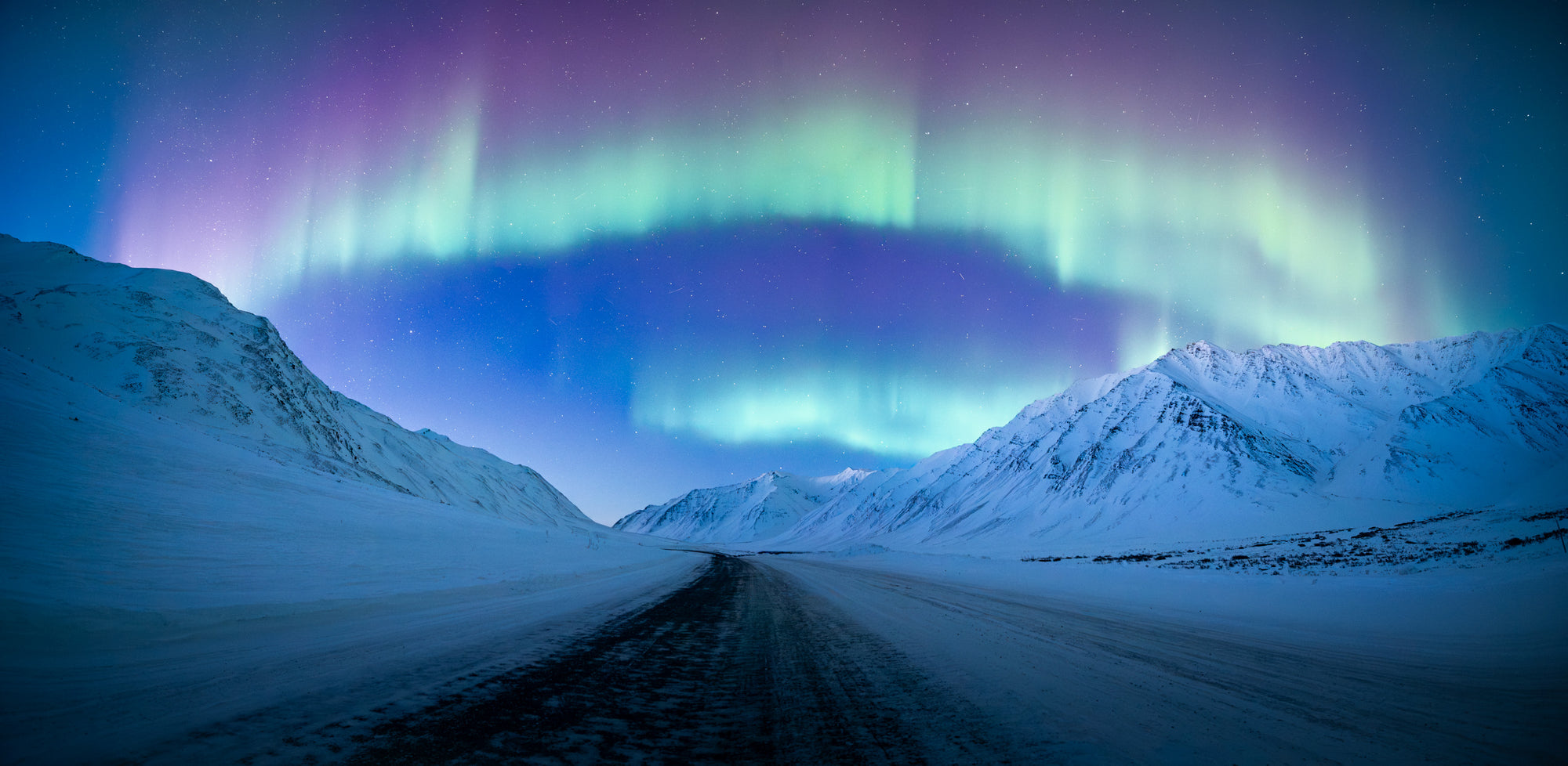 Aurora borealis arching over the Dalton Highway near Atigun Pass in Alaska, with a snow-covered road leading through Arctic mountains at night.