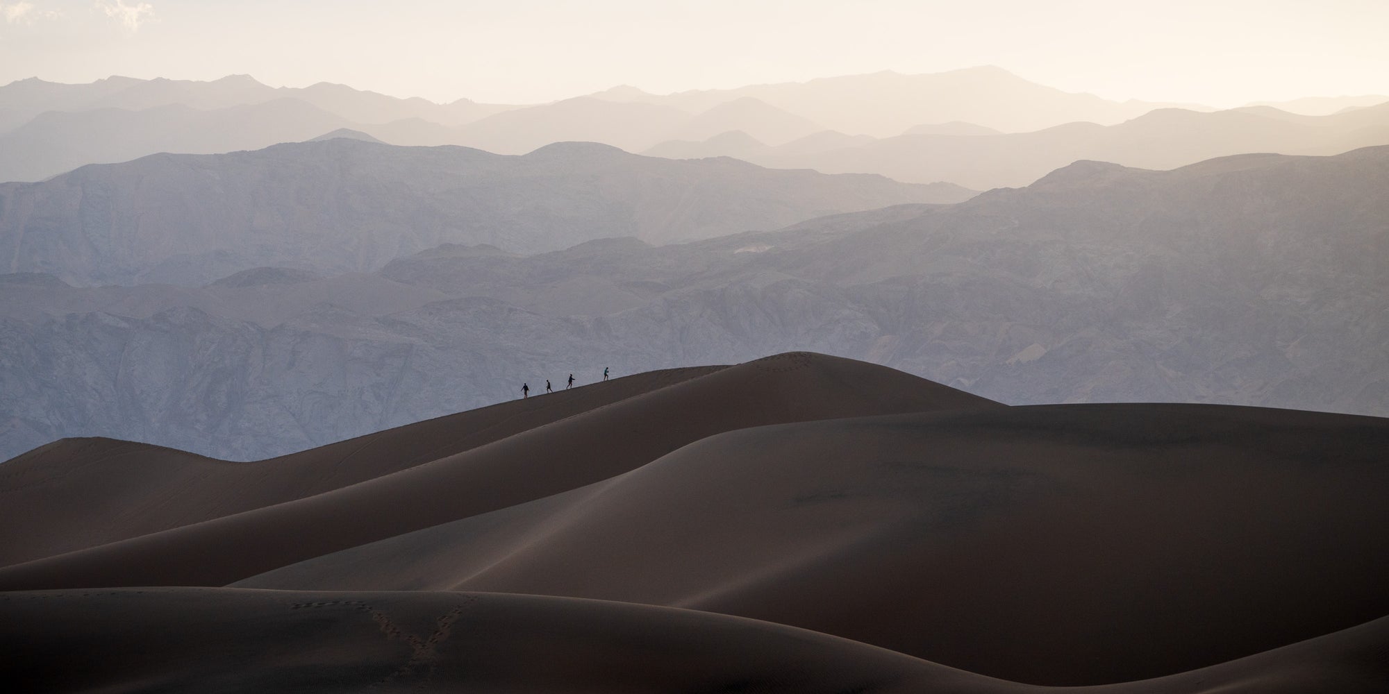 Layered sand dunes in Death Valley National Park with distant glowing mountains, showing small hikers silhouetted along a dune ridge for scale.