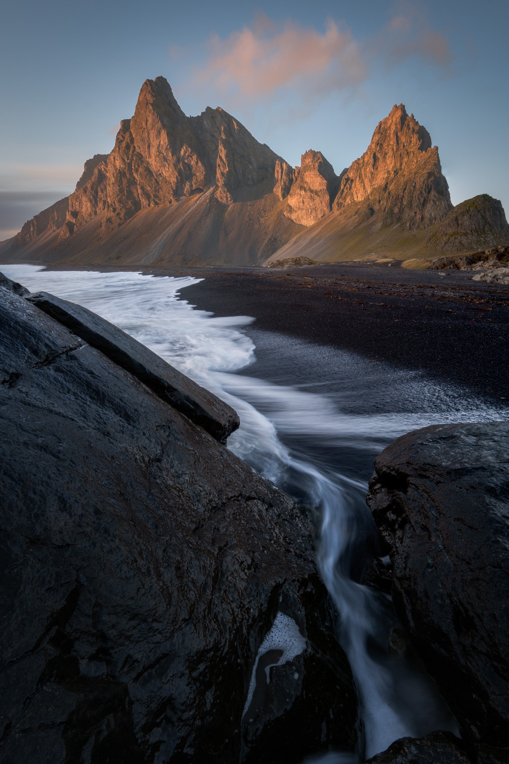 Black sand beach on Iceland’s southern coast at Eystrahorn, with waves flowing through dark volcanic rock toward jagged mountains lit by warm early light.
