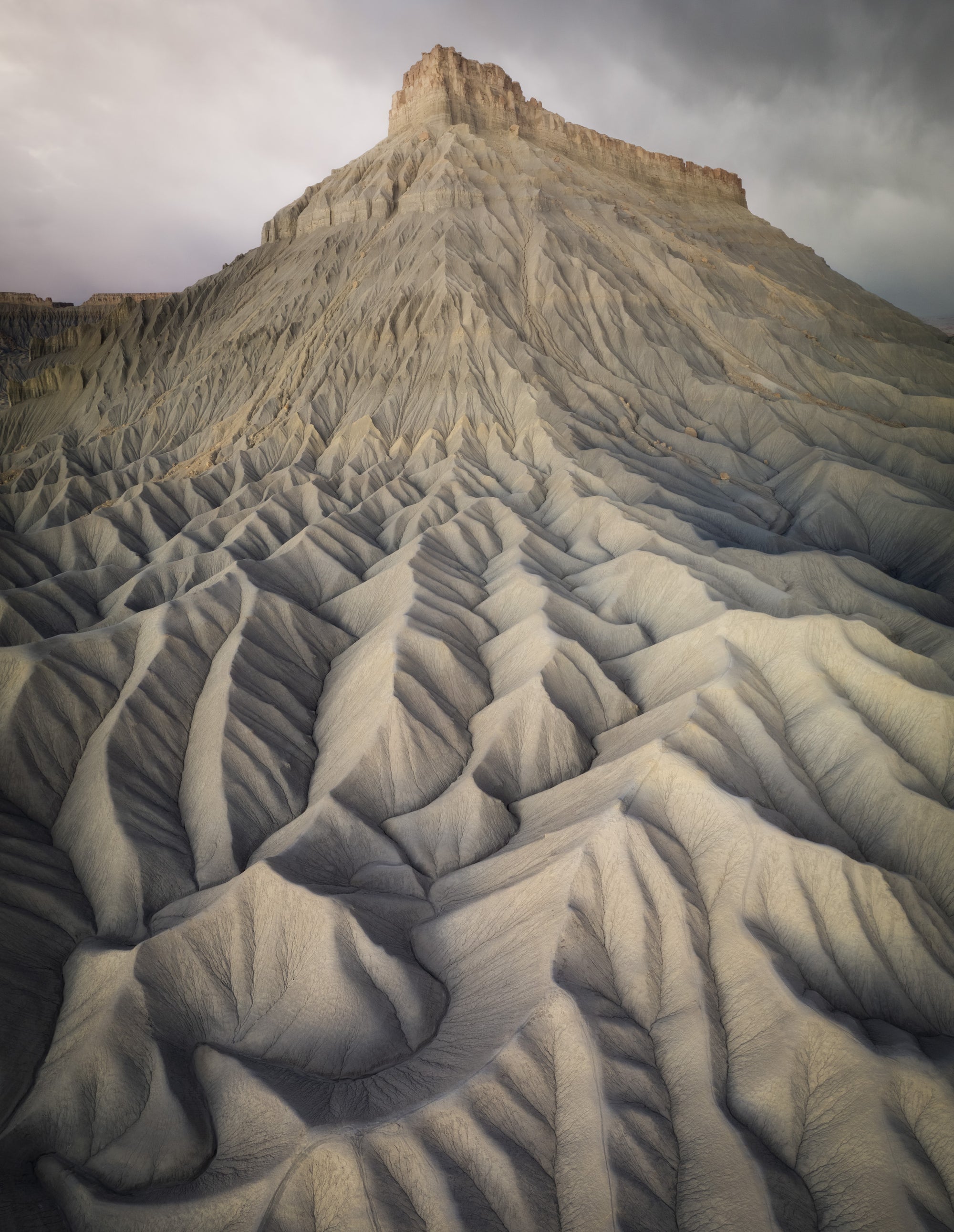 Aerial view of Factory Butte rising above textured badlands ridges in central Utah, with eroded clay patterns and stormy clouds overhead.