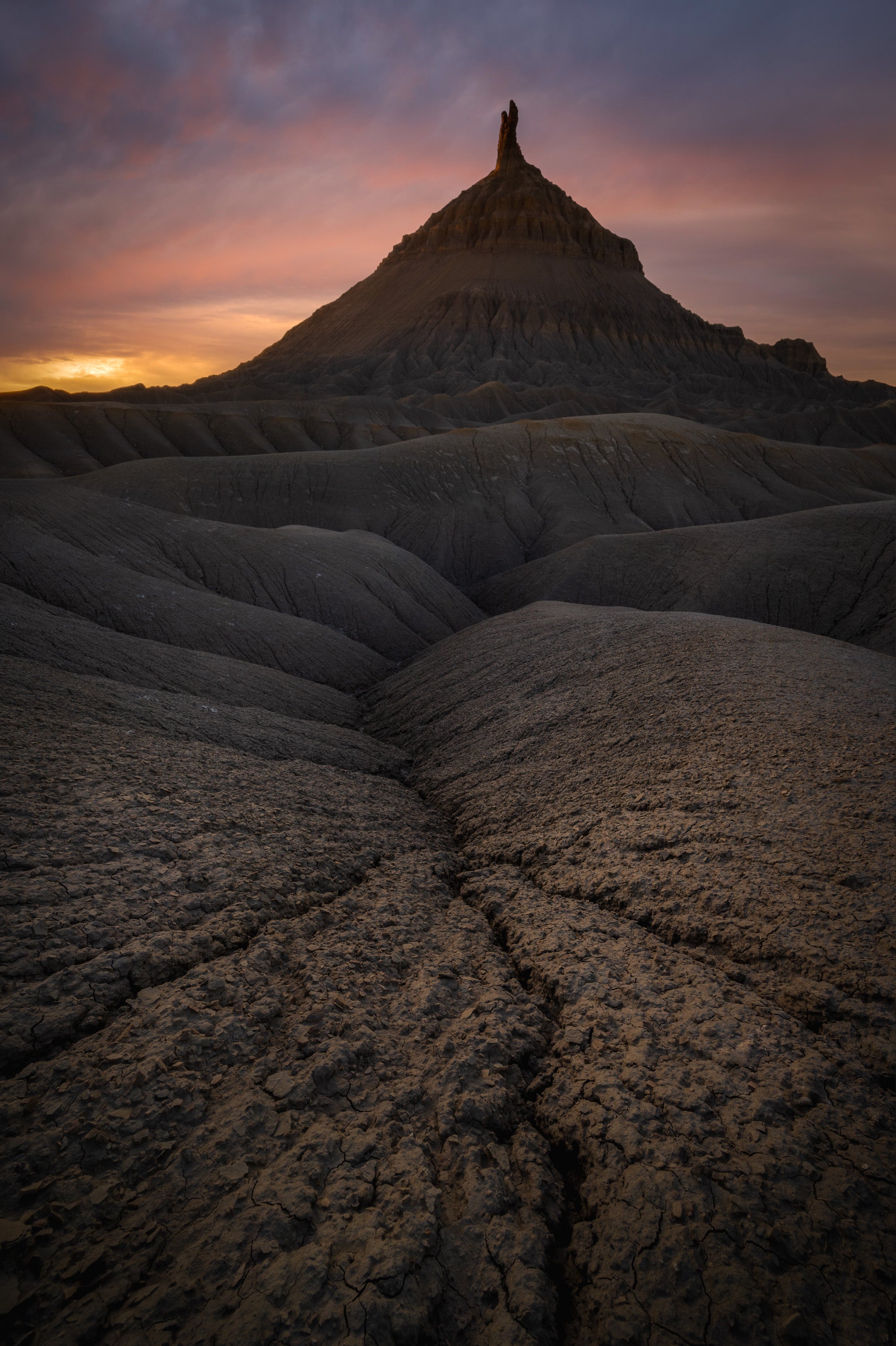 Factory Butte silhouetted at sunset in Utah’s badlands, with cracked clay textures and eroded ridges leading toward the butte beneath colorful evening clouds.