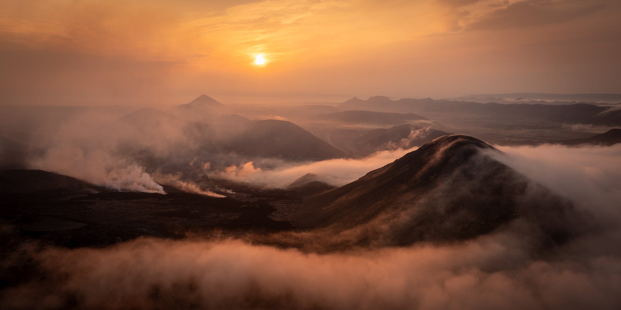 Sunrise over Iceland’s Reykjanes Peninsula during the Fagradalsfjall eruption, with volcanic smoke and low clouds drifting over dark ridges and glowing orange light near the horizon.