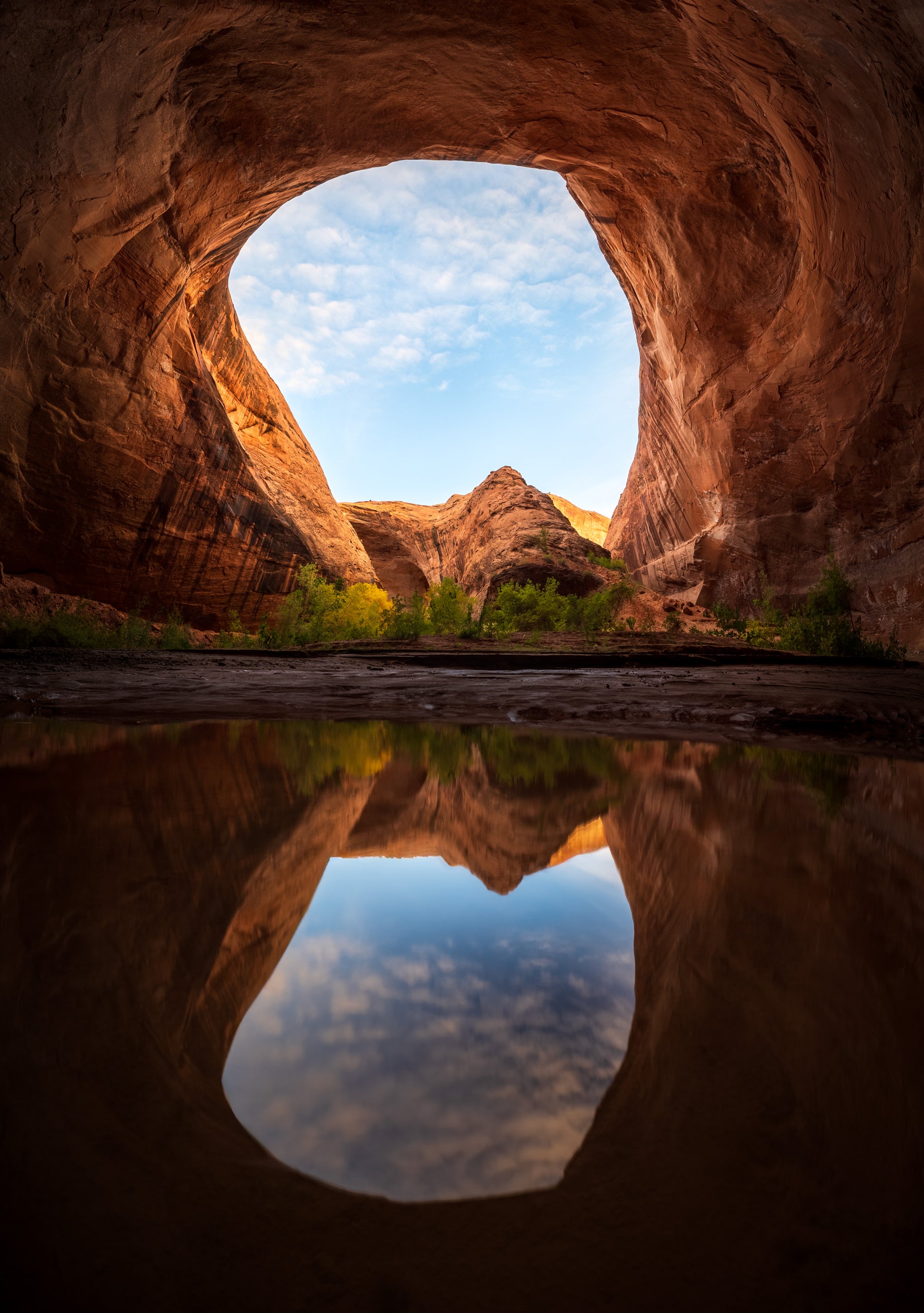 Large sandstone arch reflected in still water at sunrise in Grand Staircase–Escalante, Utah, with glowing canyon walls and blue sky framed by rock