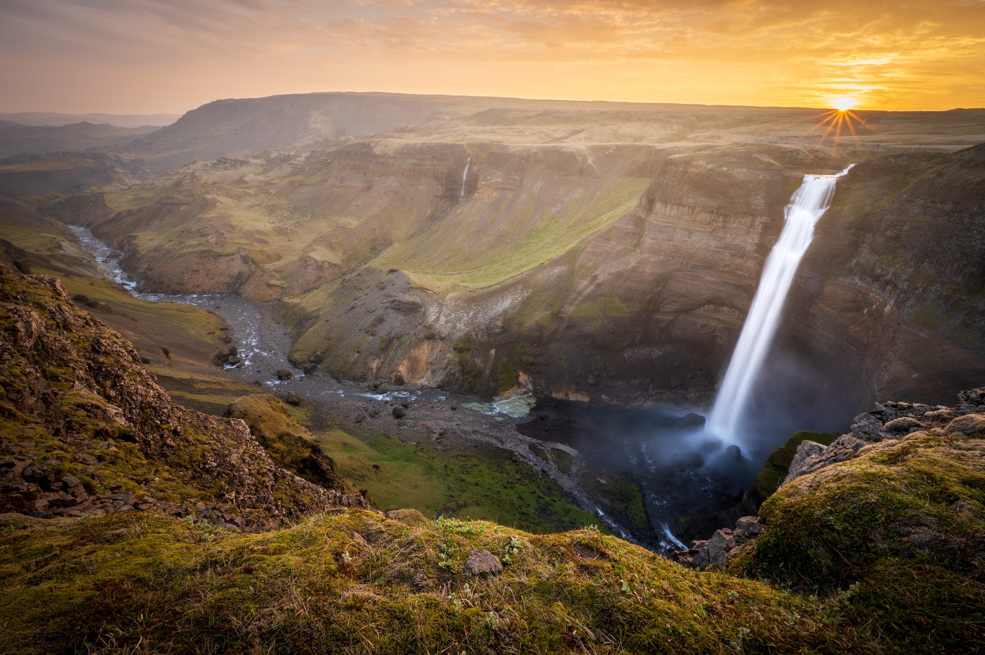Háifoss waterfall plunging into a wide Icelandic canyon under golden midnight sun light, with layered cliffs, green slopes, and a winding river below.