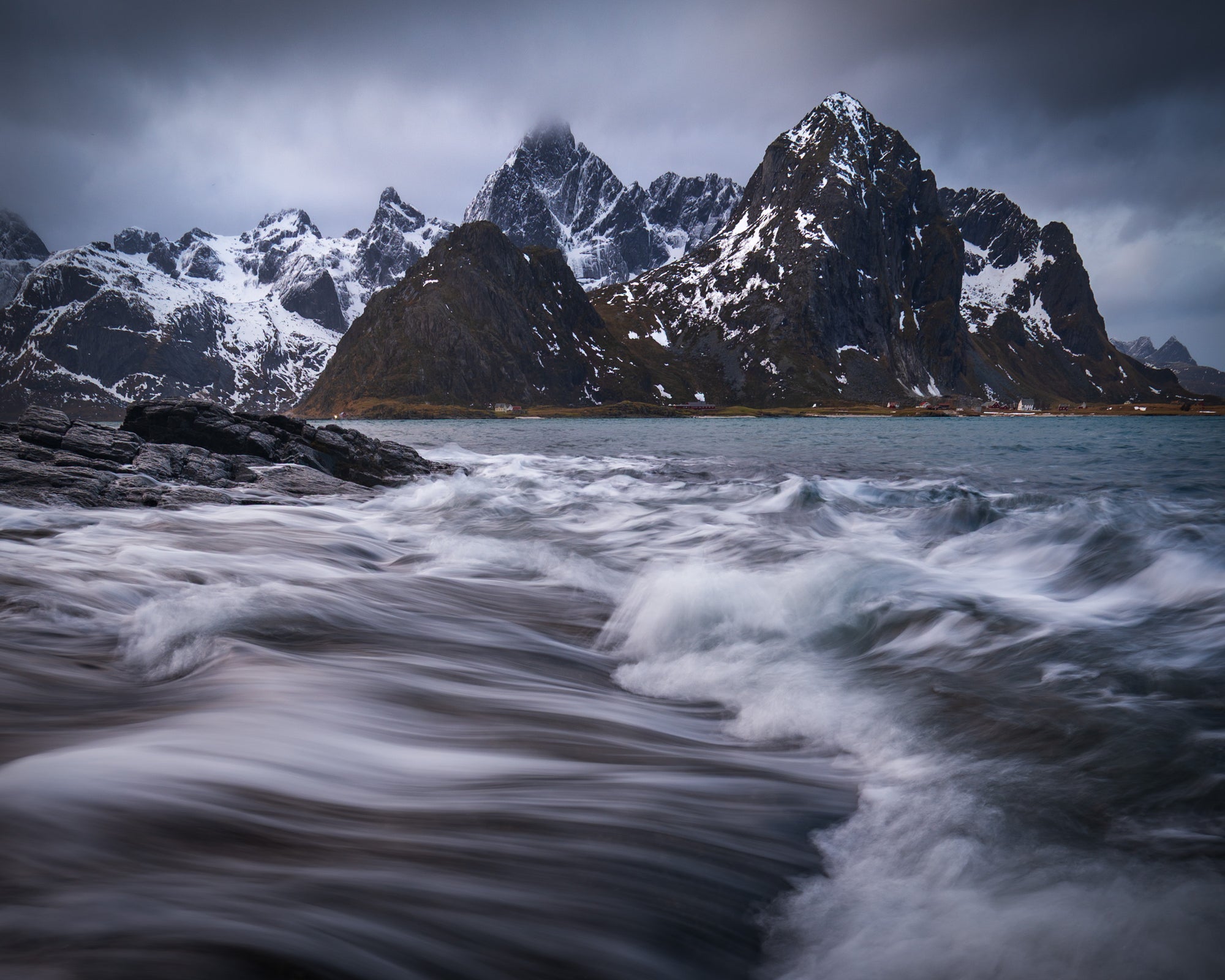 Waves crashing along the rocky coast of Norway’s Lofoten archipelago, with blurred ocean motion in the foreground and jagged snow-dusted peaks beneath stormy clouds.