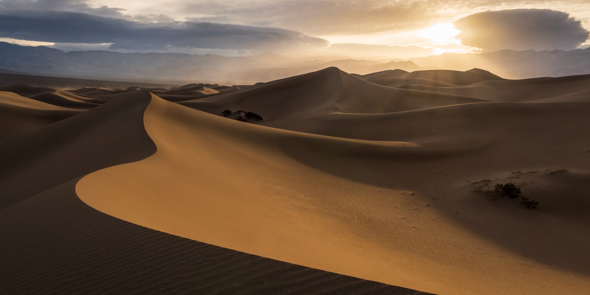 Golden sand dunes at sunset in Death Valley’s Mesquite Flat Sand Dunes, with sweeping curves leading toward distant dunes and mountains.