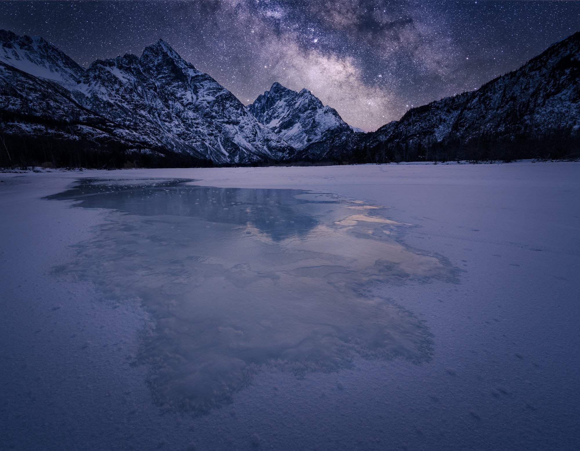 Milky Way over a frozen river near Eagle River, Alaska, with snow-covered Chugach mountains under a clear winter night sky and icy reflections in the foreground.