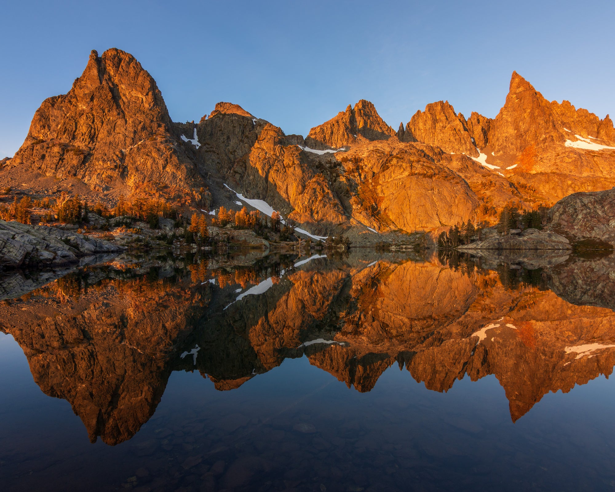 Sunrise photograph of the Minarets in the Eastern Sierra, California, glowing orange at sunrise and reflected in a calm alpine lake under a clear blue sky.