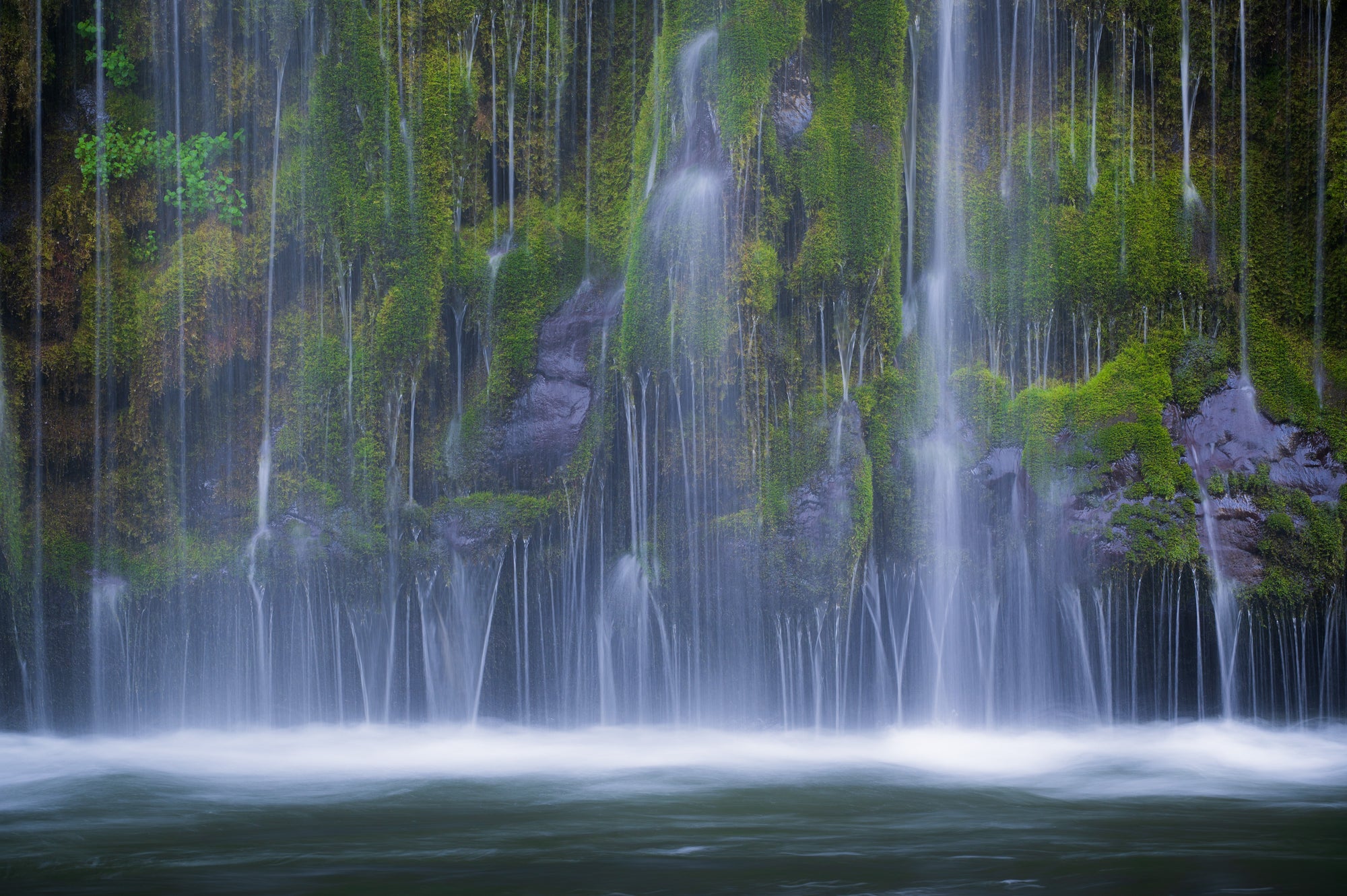 Long exposure of Mossbrae Falls in Northern California, showing silky streams of water cascading over a lush moss-covered rock wall.