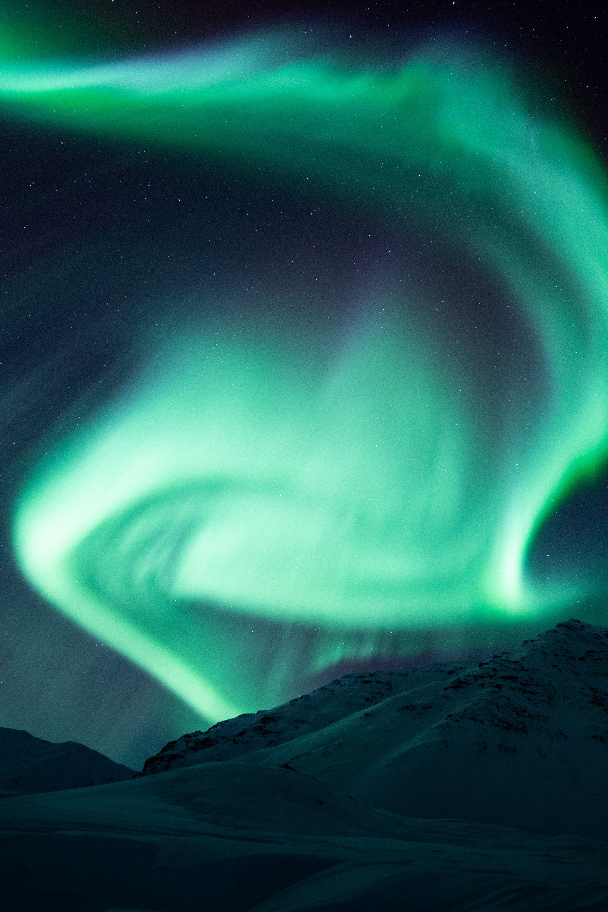 Bright green aurora borealis forming a swirling spiral above snowy mountains near Atigun Pass in Alaska on a clear Arctic night.
