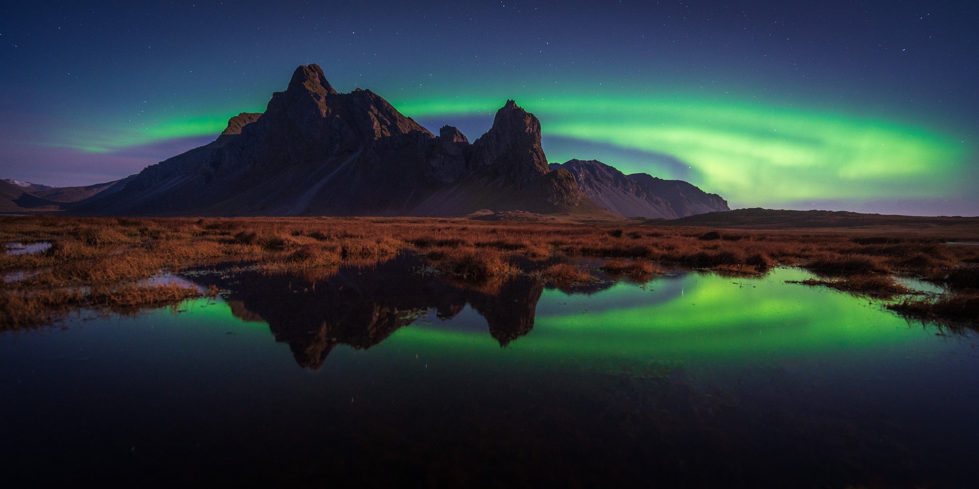 Northern lights glowing green above the Eystrahorn mountains in Iceland, reflected in still water beneath a star-filled night sky.