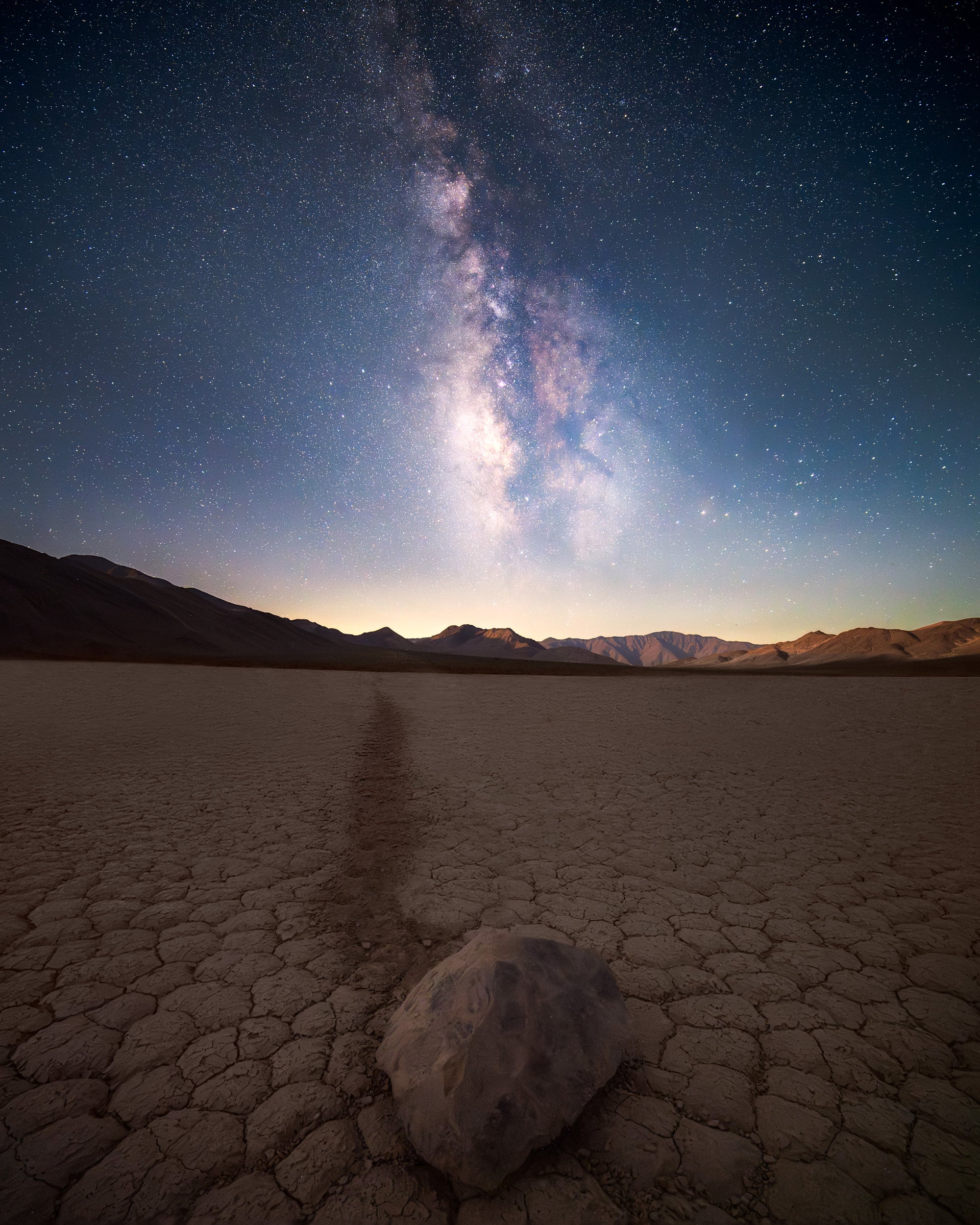 A sailing stone on Racetrack Playa in Death Valley beneath the Milky Way, with cracked desert ground and a faint rock trail leading toward distant mountains at night.