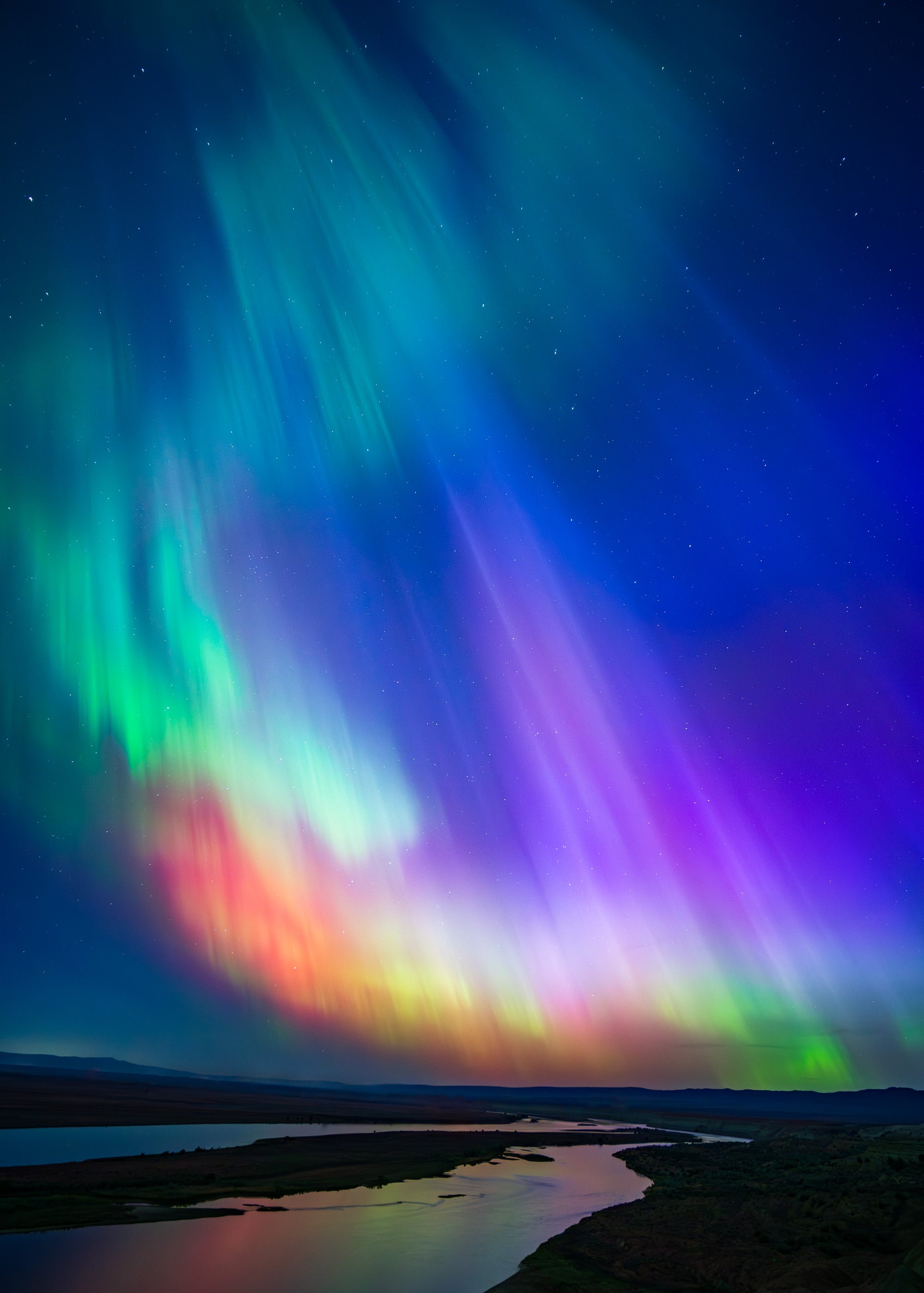 Vivid multicolored aurora borealis glowing over Eastern Washington, with green, purple, and red lights reflecting in a river during a rare geomagnetic storm.