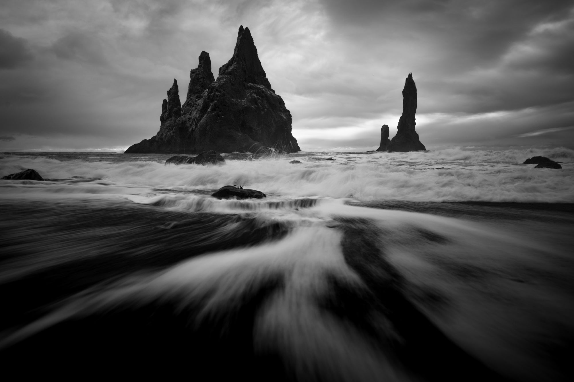 Black and white photo of Reynisfjara Beach in Iceland with blurred ocean surf leading toward the Reynisdrangar sea stacks under a dark, moody sky.