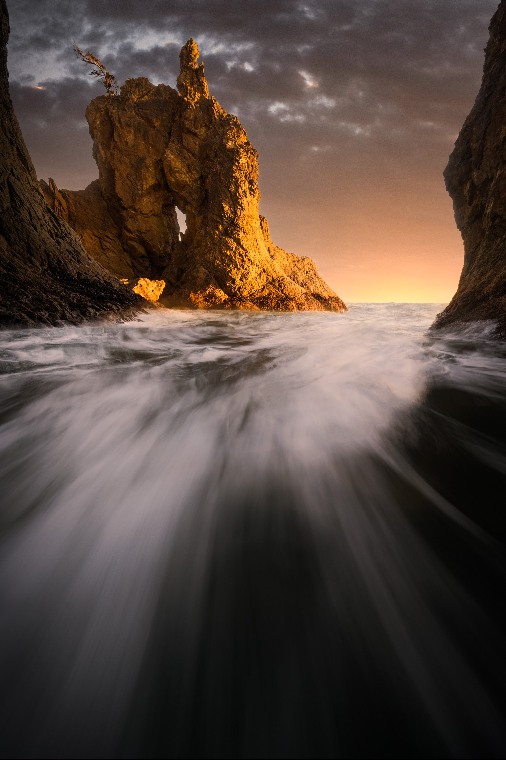 Long-exposure photograph at Rialto Beach showing waves rushing between sea stacks toward a glowing rock formation at sunset on the Olympic Peninsula.