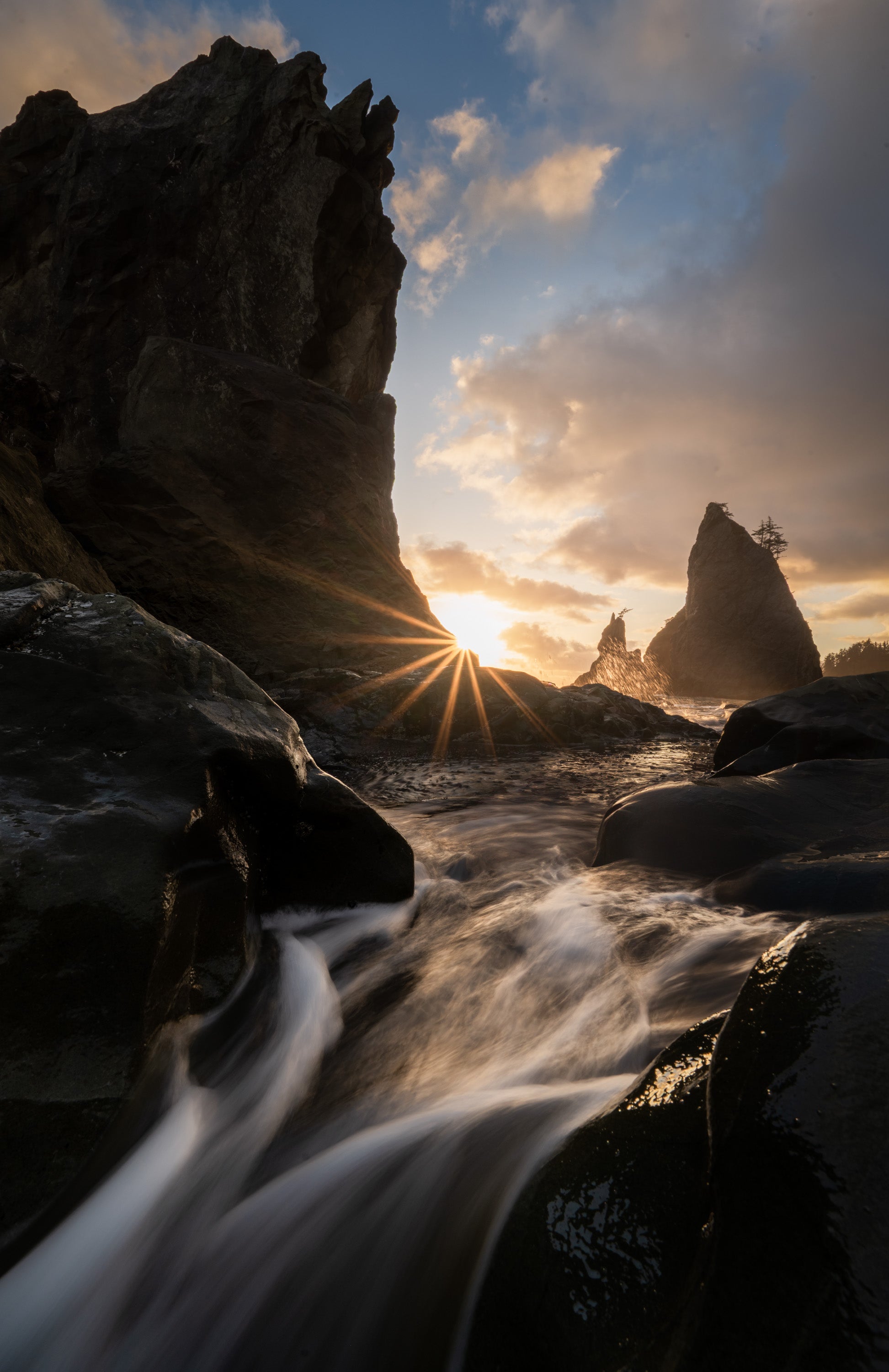 Sunburst at Rialto Beach on Washington’s Olympic Peninsula, with flowing water over dark rocks and sea stacks silhouetted along the rugged coast.
