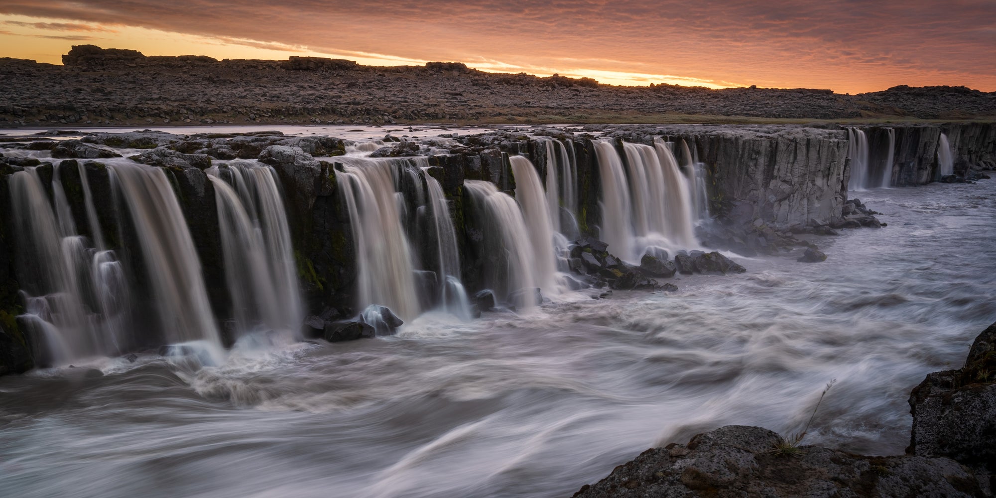 Selfoss waterfall in Iceland spilling across a wide volcanic river under midnight sun light, with fast-moving water flowing toward Dettifoss.